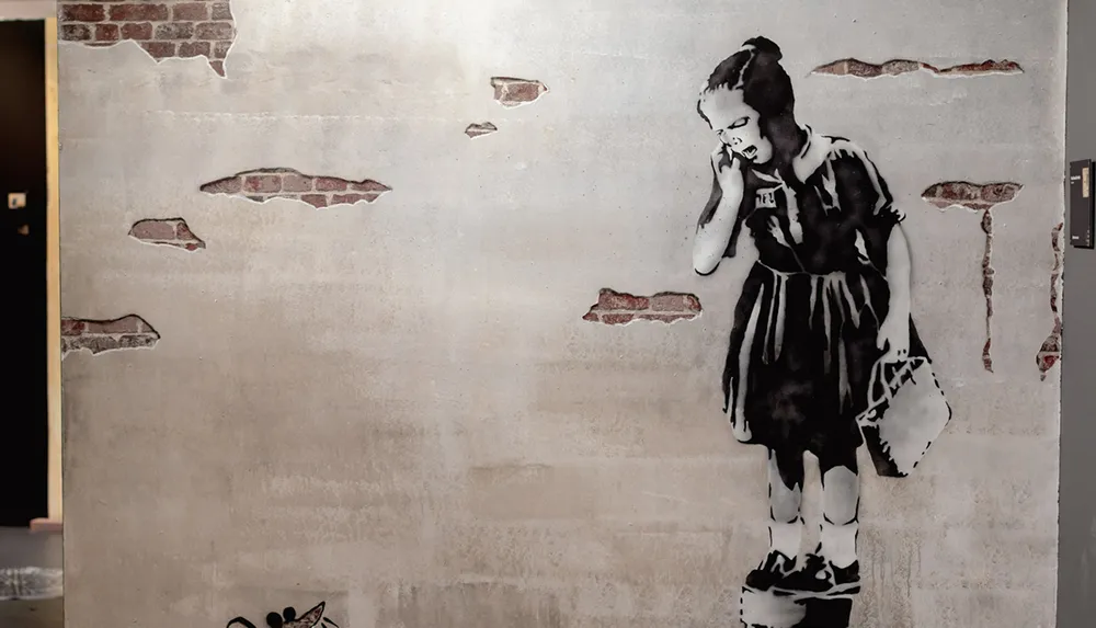 The image shows a stenciled black and white artwork of a girl in a dress crying into a phone on a wall with exposed brickwork evoking the style of street artist Banksy