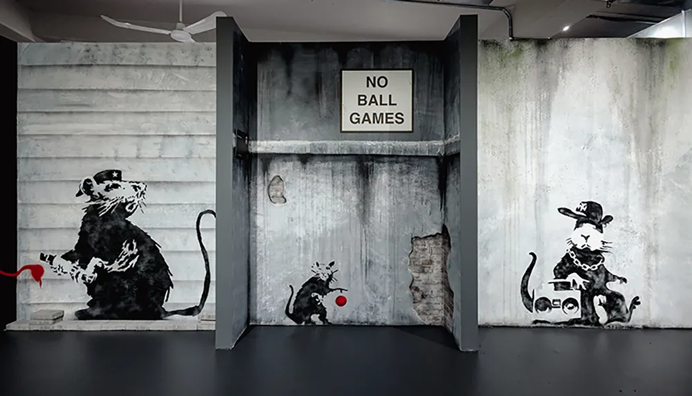 The image features a street art-style mural with stenciled rats engaging in playful activities such as bouncing a ball and driving a toy car directly beneath a sign that says NO BALL GAMES