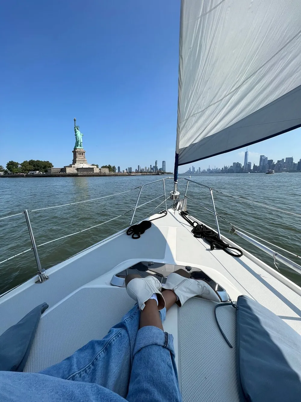 A person is relaxing on a sailboat with their feet up enjoying a clear day with a view of the Statue of Liberty and the New York City skyline in the background