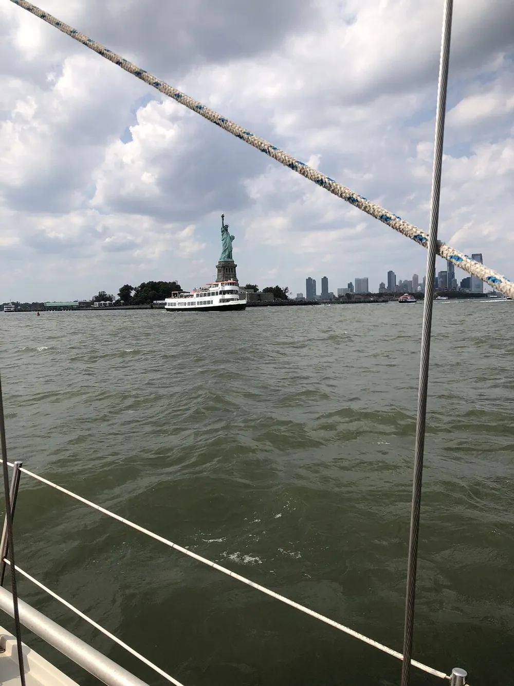 The image shows a view from a boat featuring the Statue of Liberty and a ferry with the New York City skyline in the background framed by ropes from the boats rigging