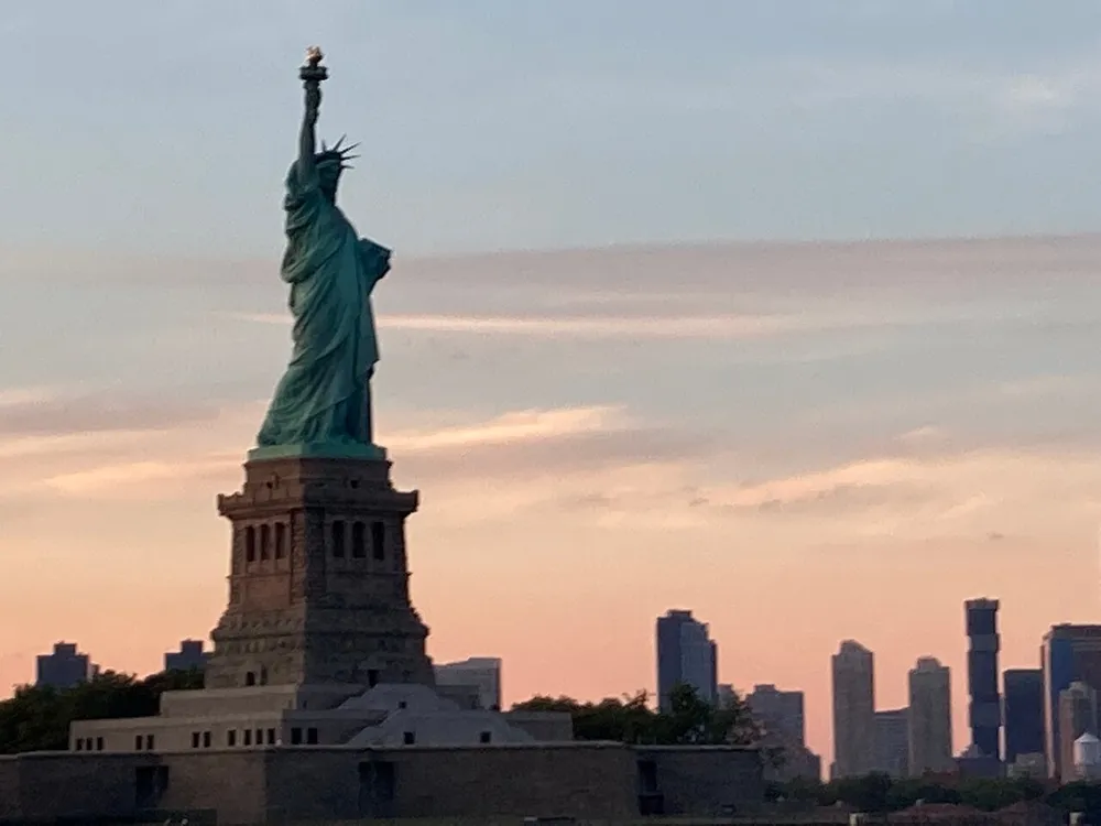 The image shows the Statue of Liberty silhouetted against a dusky sky with the outlines of city skyscrapers in the background