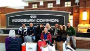 A group of smiling people with shopping bags are posing in front of the Woodbury Common Premium Outlets sign.