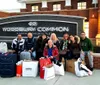 A group of people is posing with shopping bags and luggage in front of the Woodbury Common Premium Outlet sign seemingly happy after a shopping spree