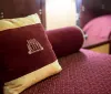 The image shows a plush maroon and gold pillow with the words Radio City Apartments NYC embroidered on it resting on a maroon patterned bedspread