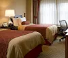 This image depicts a neatly arranged hotel room with a large bed adorned with white linens and a red decorative runner complemented by bedside furniture and artwork on the walls