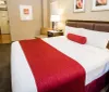 This image depicts a neatly arranged hotel room with a large bed adorned with white linens and a red decorative runner complemented by bedside furniture and artwork on the walls