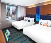 The image shows a modern hotel room with a neatly made bed vibrant decor and a view of city buildings through the window