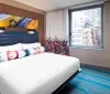 The image shows a modern hotel room with a neatly made bed vibrant decor and a view of city buildings through the window