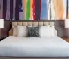 The image shows a neatly made bed with a tufted headboard flanked by two table lamps against a backdrop of a large colorful abstract painting
