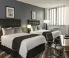 The image shows a well-lit modern hotel room with two neatly made beds a desk with a chair striped carpet dark grey walls and framed artwork