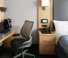 The image shows a neatly organized hotel room with a large bed a wall-mounted TV a desk with a chair and sheer curtains partially drawn over a window