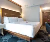The image shows a neatly arranged hotel room with a large bed a work desk with a chair and modern decor
