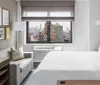 The image shows a modern hotel room with a large bed a couch a television and a window offering a view of a city skyline