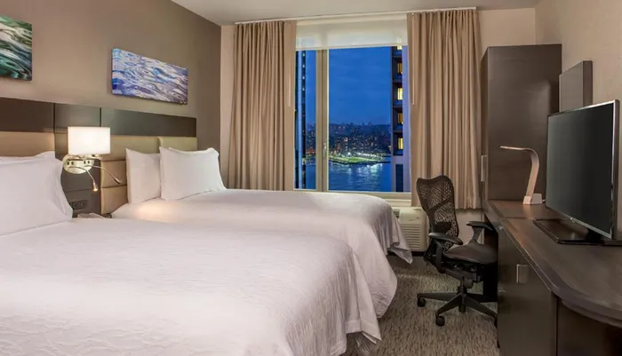 A modern hotel room with two beds features a large window offering a nighttime cityscape view complemented by tasteful decor and a work desk with a television