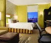 This image features a modern hotel room with a cozy bed a seating area a workspace and a large window that offers a stunning night view of a city skyline