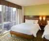 The image shows a modern hotel room with two beds simple decor and a large window offering a view of a bustling cityscape