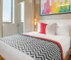 The image shows a neatly arranged hotel room with a large bed featuring a geometric-patterned blanket accented with a red throw pillow and a colorful cityscape artwork above the headboard