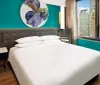 The image shows a modern bedroom with a large bed with teal walls colorful artwork and a view of an urban environment through the window