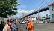 Three people wearing bicycle helmets are posing for a photo with a backdrop of the Brooklyn Bridge and New York City skyline.