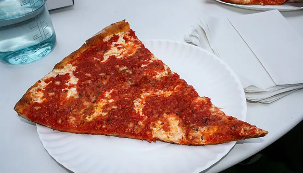 A large slice of cheese pizza with a vibrant red sauce is resting on a white paper plate