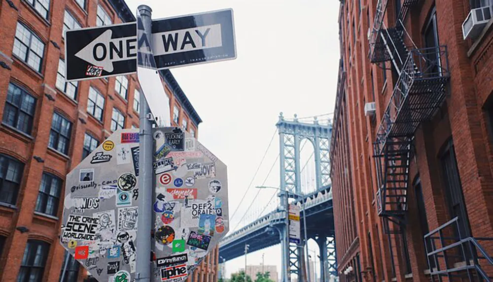 A street sign pole covered in various stickers stands before a backdrop of red brick buildings and a steel bridge under a cloudy sky