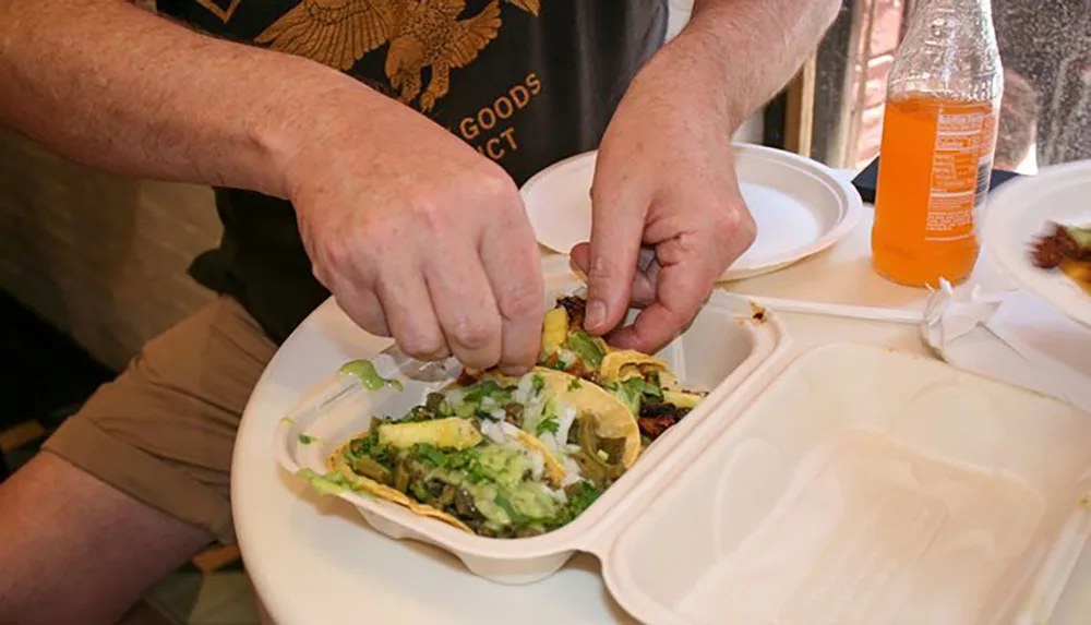 A person is squeezing a lime over tacos in a foam takeout container with a bottle of orange soda nearby