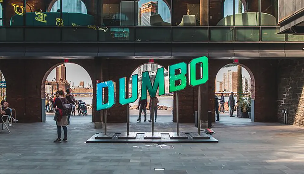 The image shows a large colorful DUMBO sign in a pedestrian area between two sections of a building with people walking about and engaging in various activities