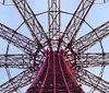 The image shows a close-up worms-eye view of a metallic structure possibly a Ferris wheel or a tower with intricate steel beams illuminated by pink lighting against a twilight sky