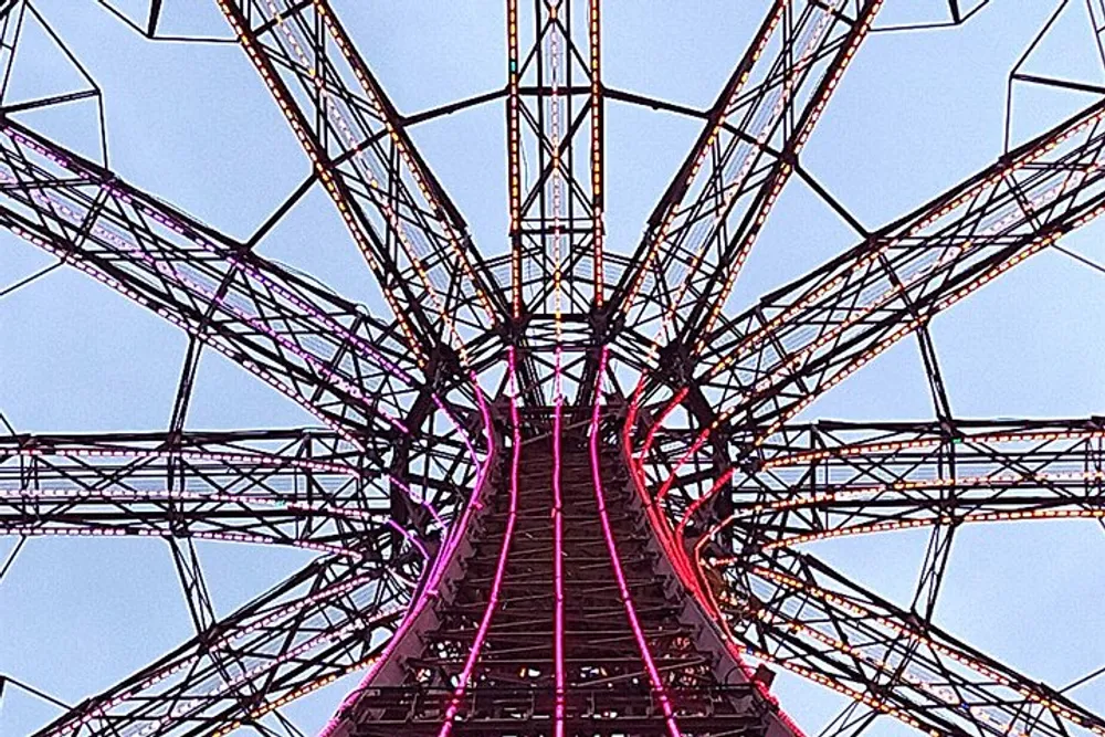 The image shows a close-up worms-eye view of a metallic structure possibly a Ferris wheel or a tower with intricate steel beams illuminated by pink lighting against a twilight sky