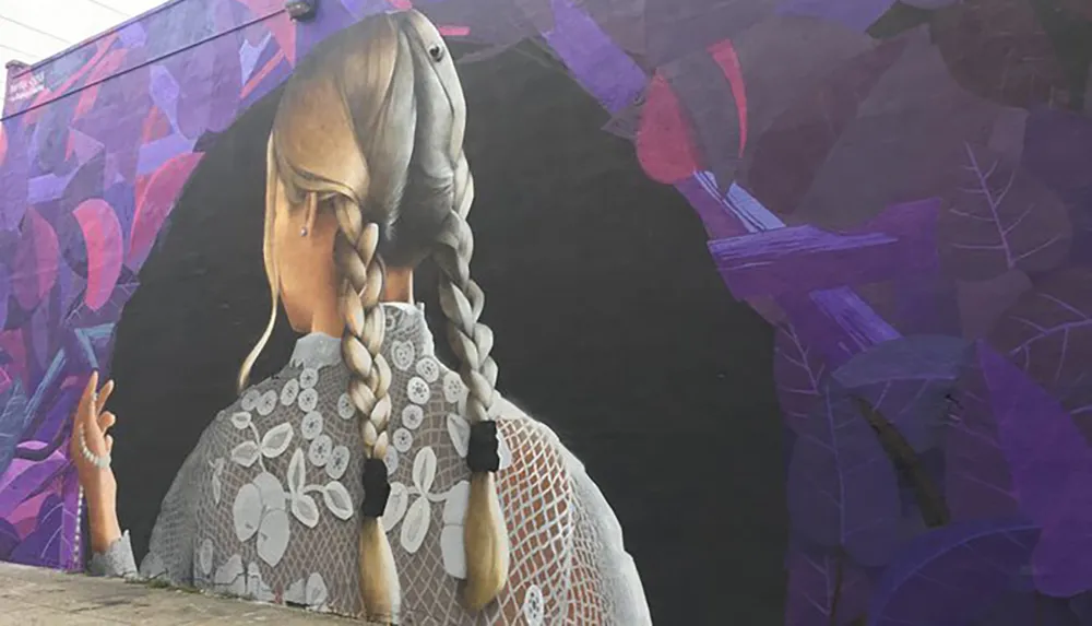 The image shows a large-scale mural of a womans back with detailed braids and a patterned outfit set against a background of abstract purple foliage