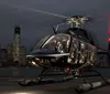 A helicopter with illuminated cabin lights is parked on a helipad at dusk with a city skyline in the background
