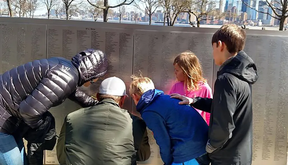 A group of people is gathered around a memorial wall engrossed in reading and pointing at the inscriptions