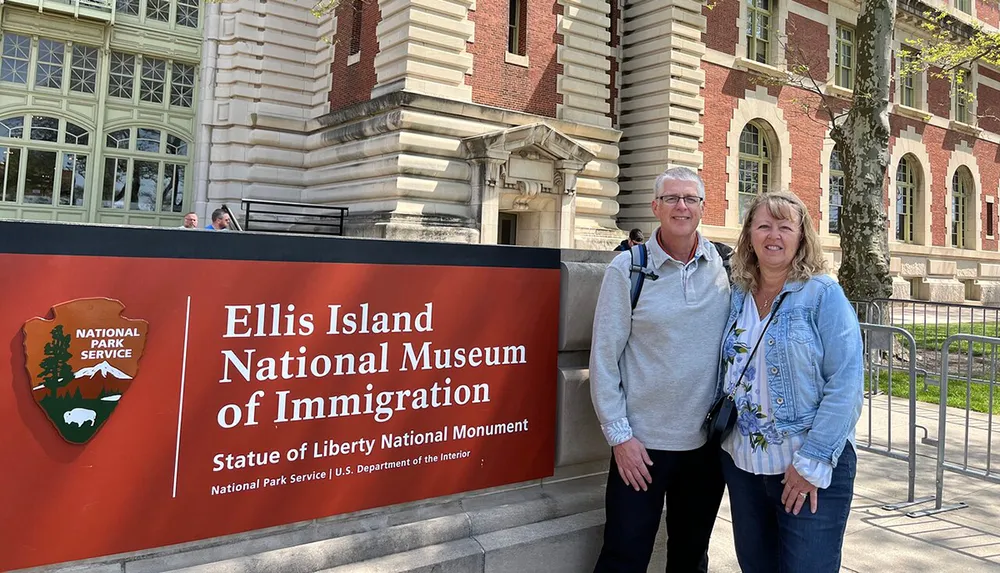 Two smiling individuals are posing in front of the Ellis Island National Museum of Immigration sign with the historic red-brick building in the background