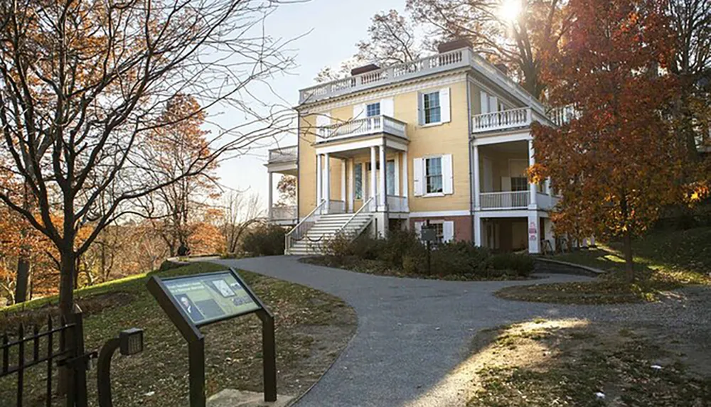 The image shows a stately yellow house with white columns and balconies situated on a leaf-covered lawn with bare trees and an information plaque in the foreground in the gentle light of either sunrise or sunset