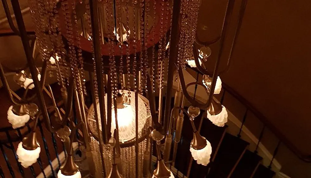 The image shows a richly detailed chandelier with numerous crystal-like beads and glass lampshades overlooking a spiral staircase cast in warm ambient lighting