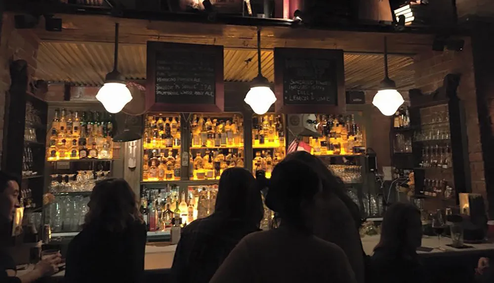 The image shows patrons at a cozy bar with a warmly-lit shelf of bottles and hanging pendant lights and chalkboard menus visible in the background