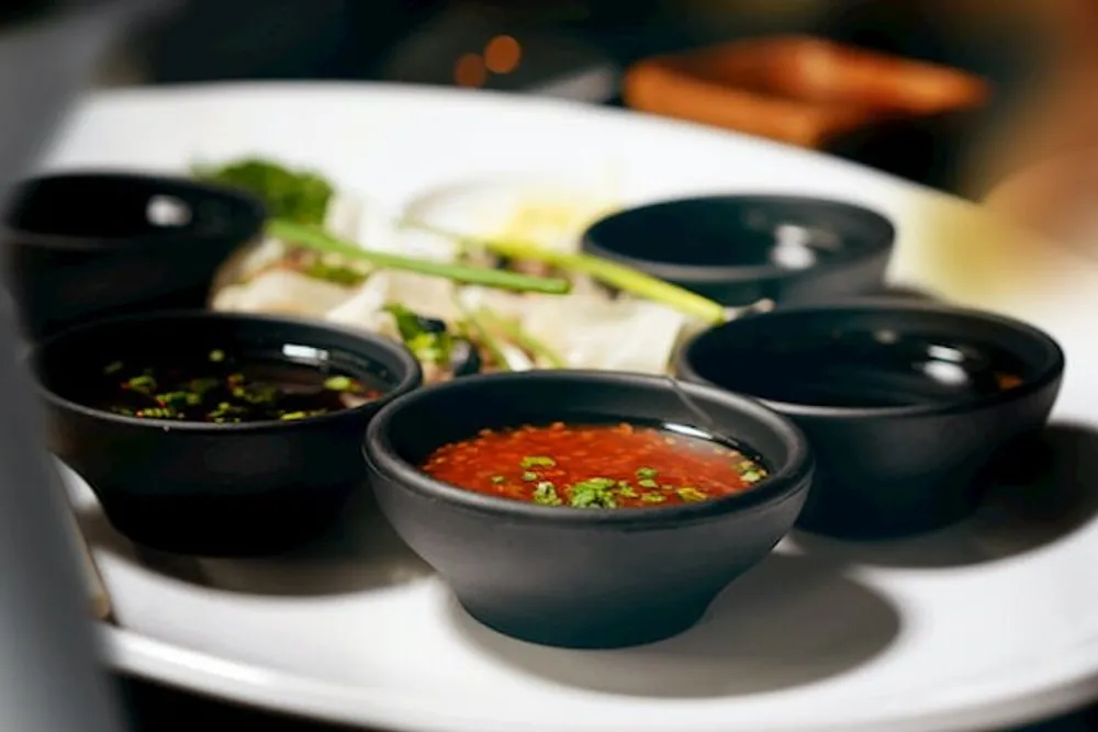 A white plate hosts several small black bowls with various sauces and some garnishes