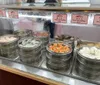 The image displays a variety of Chinese dim sum dishes in traditional steaming baskets set on a counter for sale with price tags for each dish visible in the background