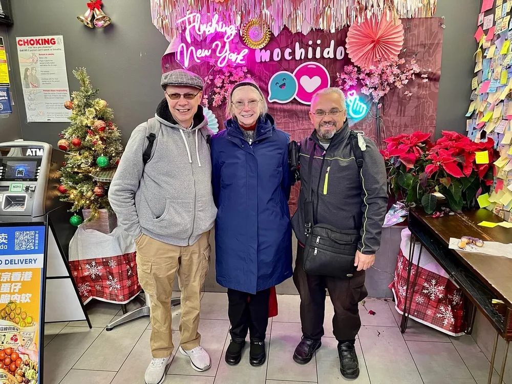 Three people are smiling for a photo inside a festive-looking shop decorated with Christmas and floral elements including a sign that reads Flushing New York mochido