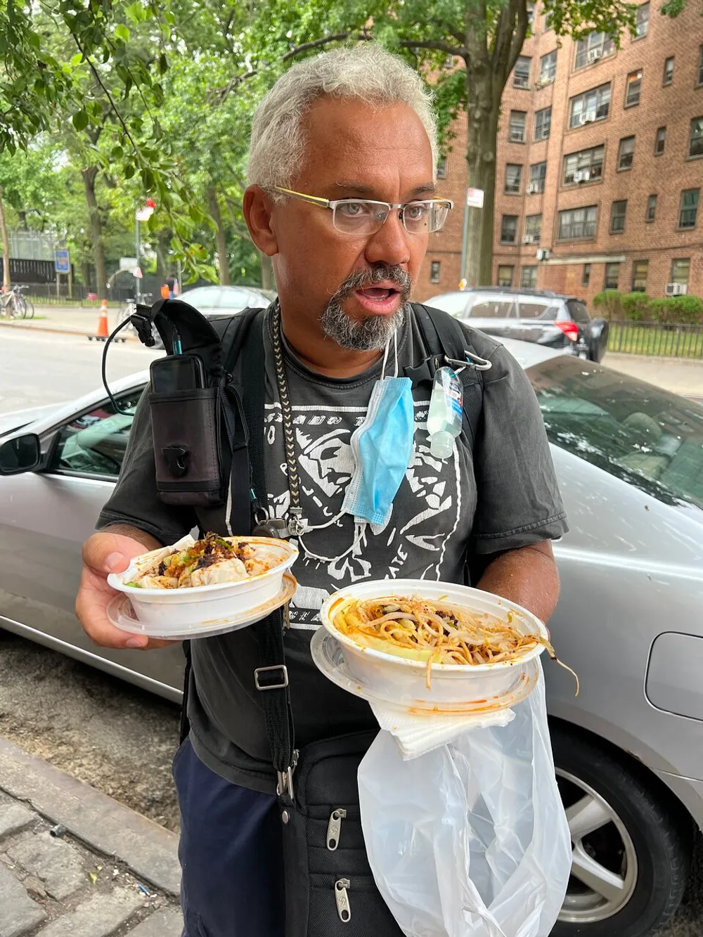 A man with glasses and a face mask dangling from his ear looks surprised while holding two plates of food outdoors
