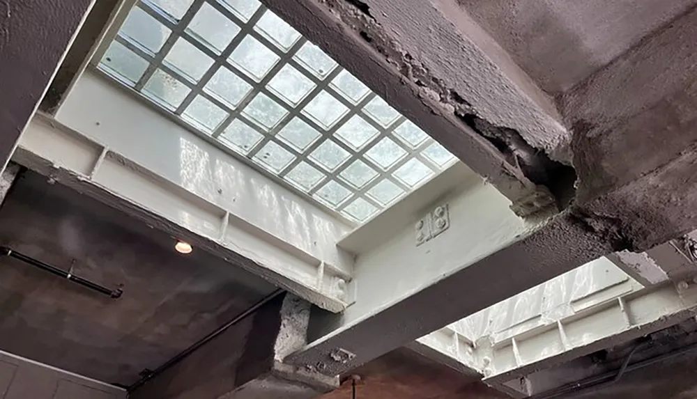 The image shows a damaged concrete ceiling with exposed reinforcement at the point of collapse below what appears to be a glass block window