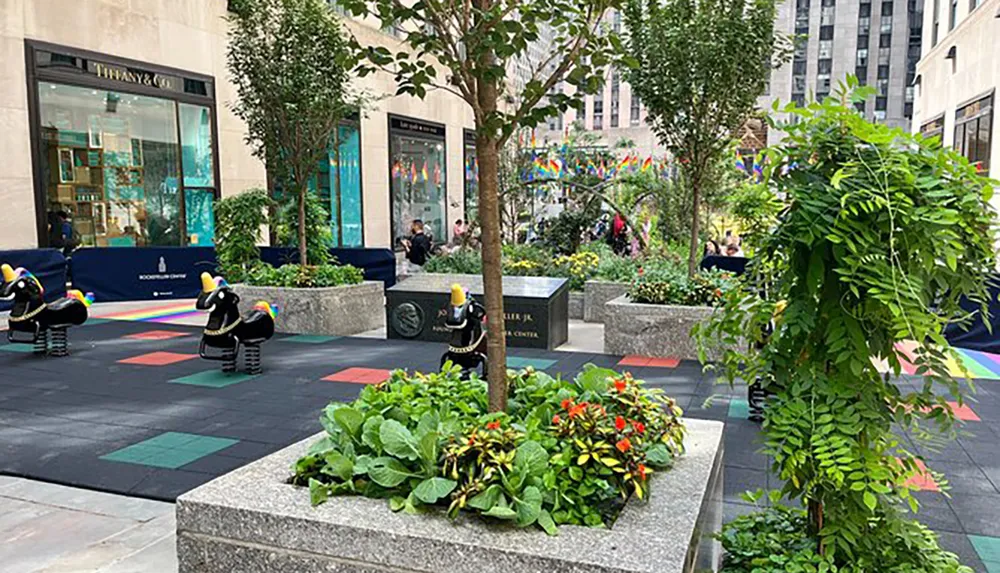 The image shows an urban plaza with playful sculptures colorful pavement plants benches and people in the background indicating a lively outdoor public space