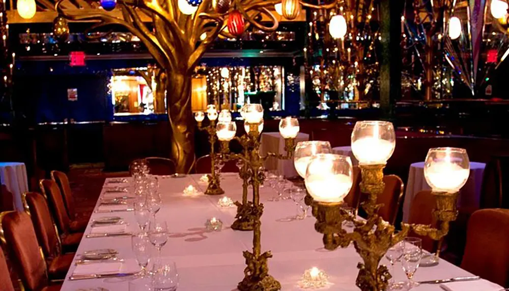 The image shows an opulent dining setting with a long table adorned with candles and glassware set within a room featuring a decorative golden tree and mirrored walls creating a luxurious ambiance