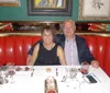 A couple is sitting at a well-set table with red upholstery in a restaurant smiling at the camera with wine glasses and dessert in front of them