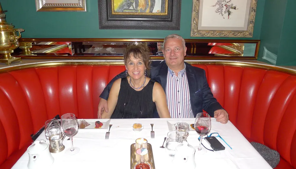 A couple is sitting at a well-set table with red upholstery in a restaurant smiling at the camera with wine glasses and dessert in front of them