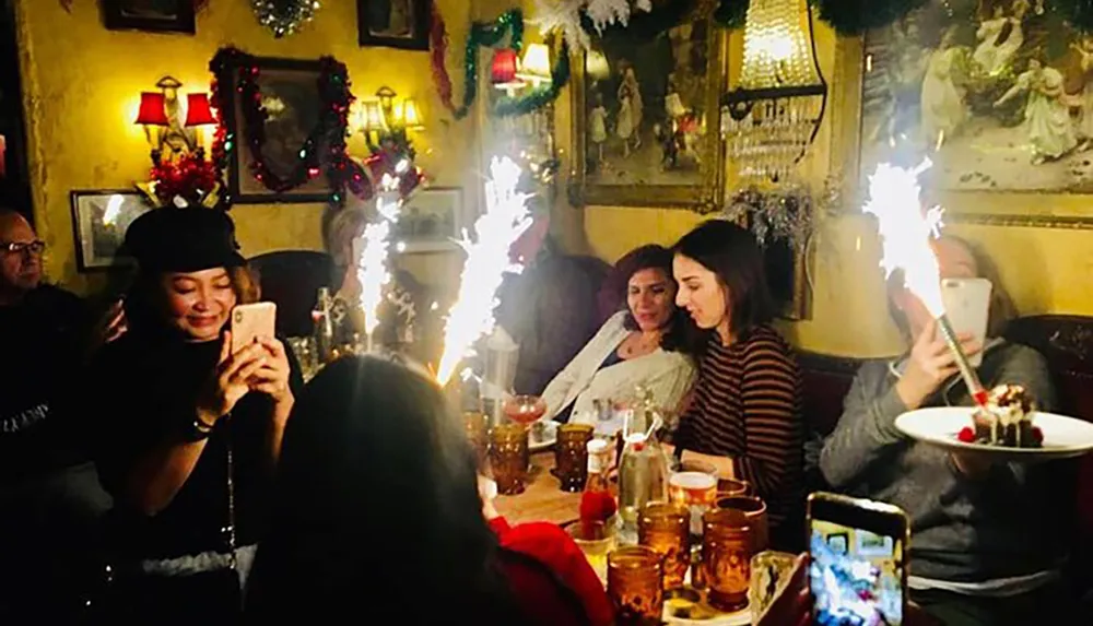 A group of people are enjoying a festive gathering in a warmly lit room with some capturing the moment with their smartphones as a dessert topped with a lit sparkler is being served