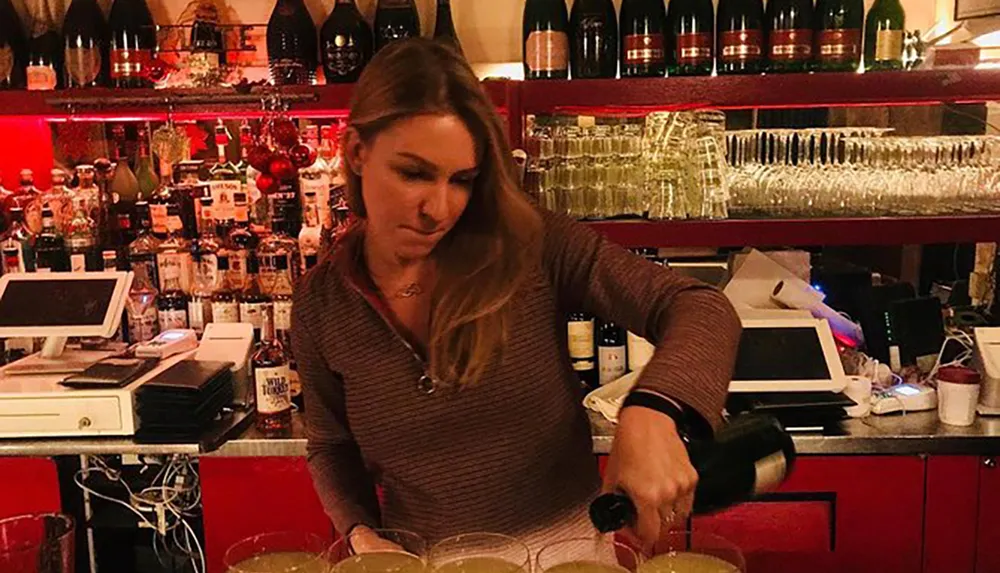 A bartender is pouring a drink at a bar with shelves stocked with various bottles in the background