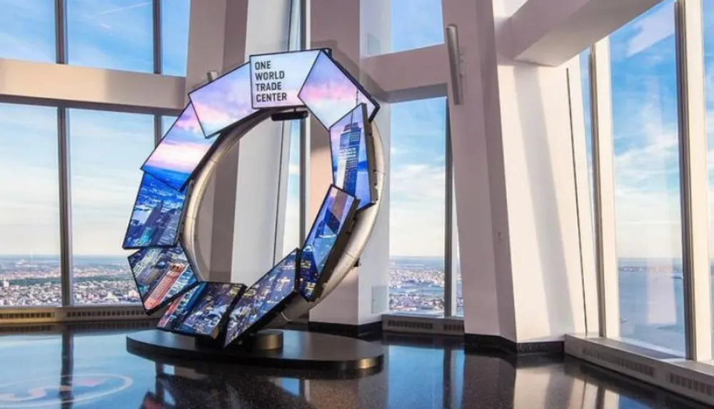 The image shows a modern interactive display designed as a ring with digital screens showing various content which is situated inside the observation area of One World Trade Center offering expansive views from large windows