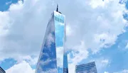 The image shows a tall skyscraper with a reflective glass facade reaching into a cloudy blue sky, surrounded by other buildings.