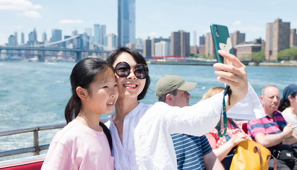 Two individuals are taking a selfie with a smartphone on a sunny day with a city skyline and a bridge in the background